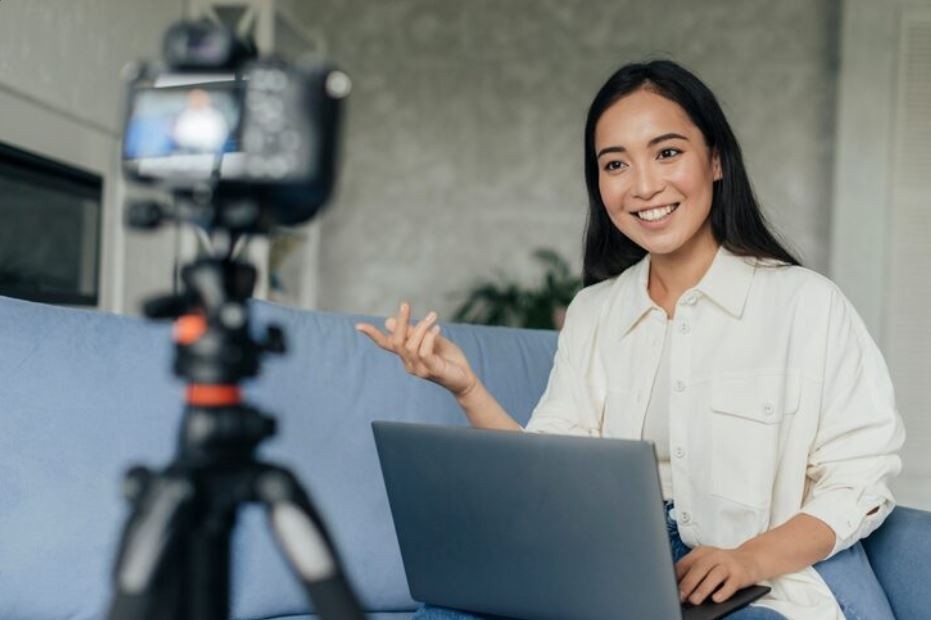 A happy woman in a white shirt sits on a sofa with a laptop, being filmed by a camera on a tripod, showcasing professional video services from a Business Videos Company in Village Of Oak Lake.