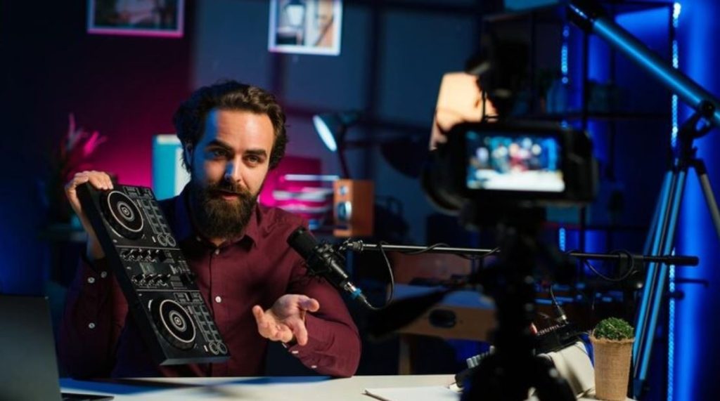 A man with a beard and a red shirt holds up a DJ controller while being filmed, showcasing a product review produced by a Business Videos Company in Sugar Land