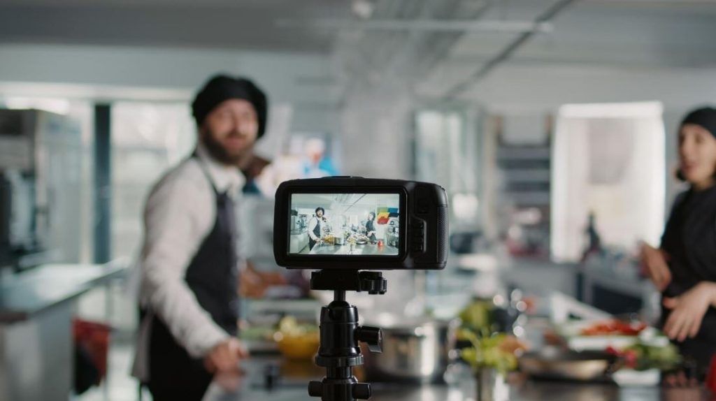 A video camera on a tripod recording two chefs in a commercial kitchen, demonstrating the types of content produced by a Business Videos Company in Park Pointe