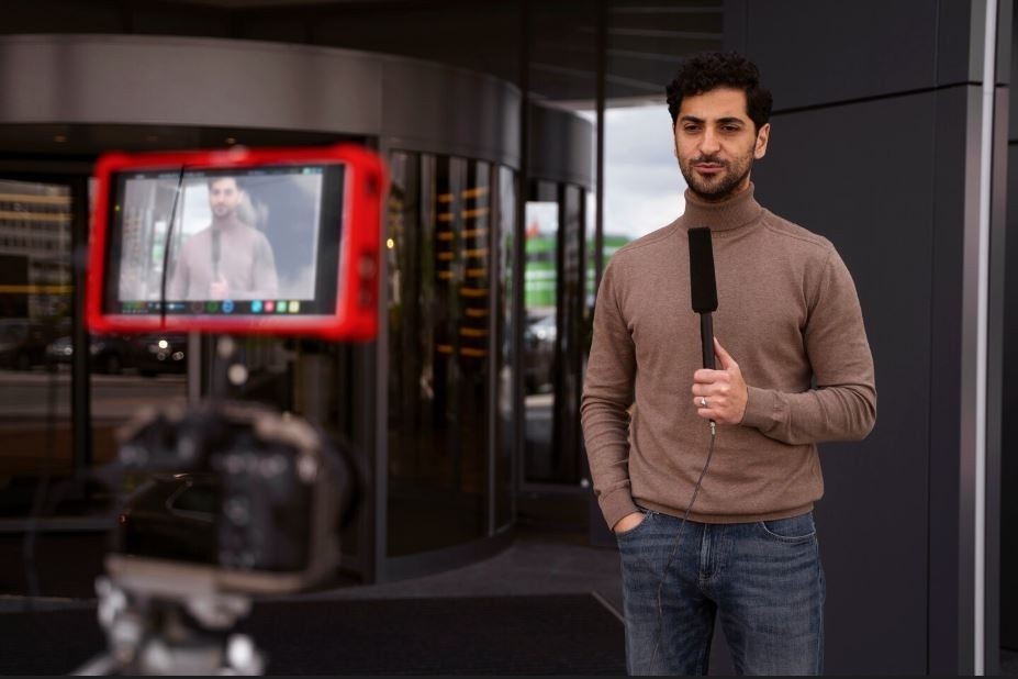 A male reporter stands with a microphone in front of a modern building, being filmed for a news-style business video by a Business Videos Company in Sugar Land