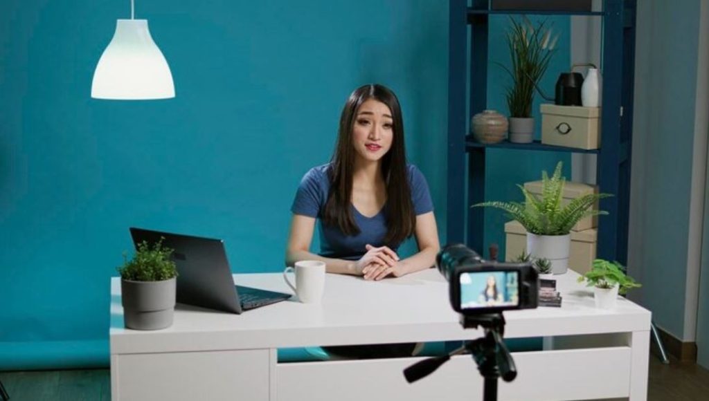 A woman sits at a desk with a laptop and plants, recording a video for her business with a camera, illustrating services from a Business Videos Company in Village Of Oak Lake.