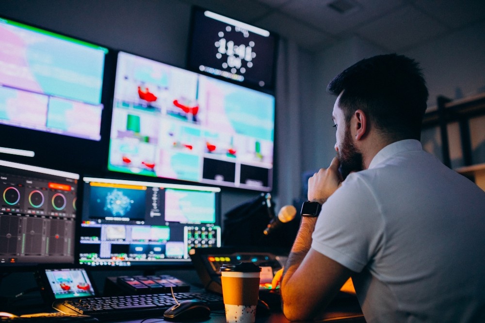 A man works at a control desk with multiple monitors, editing and monitoring a live video production, showcasing the post-production services of a Business Videos Production company in Sugar Land