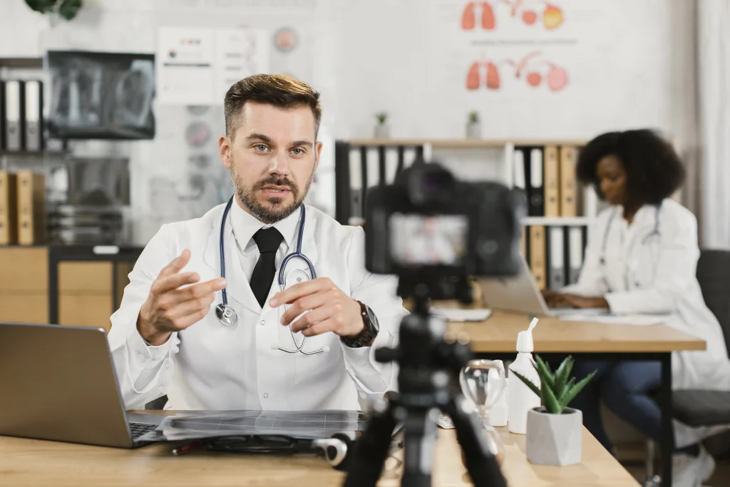 Dental videos company Sugar Land: Smiling male doctor raising finger to emphasize point while filming educational content in bright office, camera capturing engaging dental explanation.