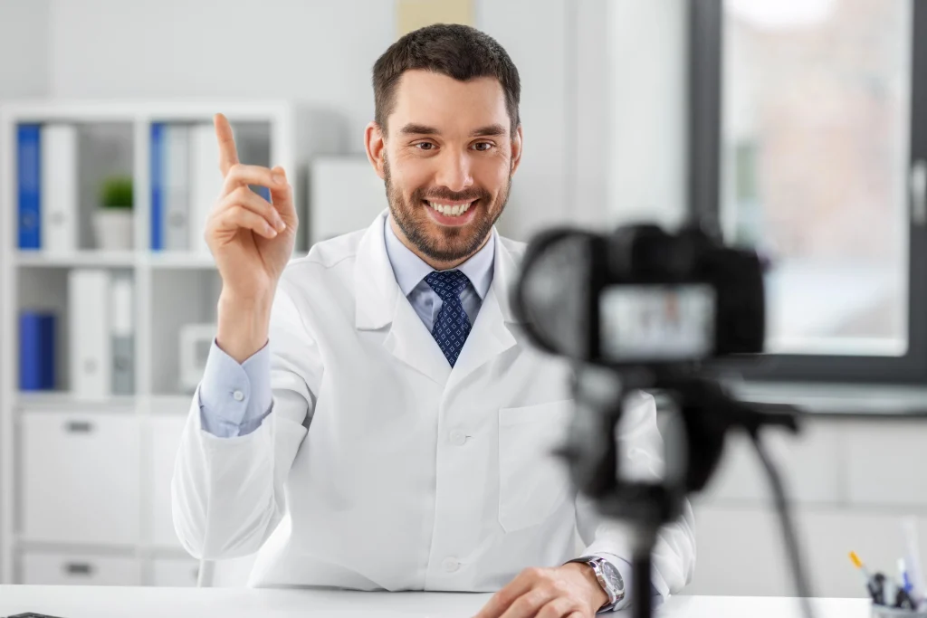 Dental videos company Sugar Land: Smiling male doctor raising finger to emphasize point while filming educational content in bright office, camera capturing engaging dental explanation.