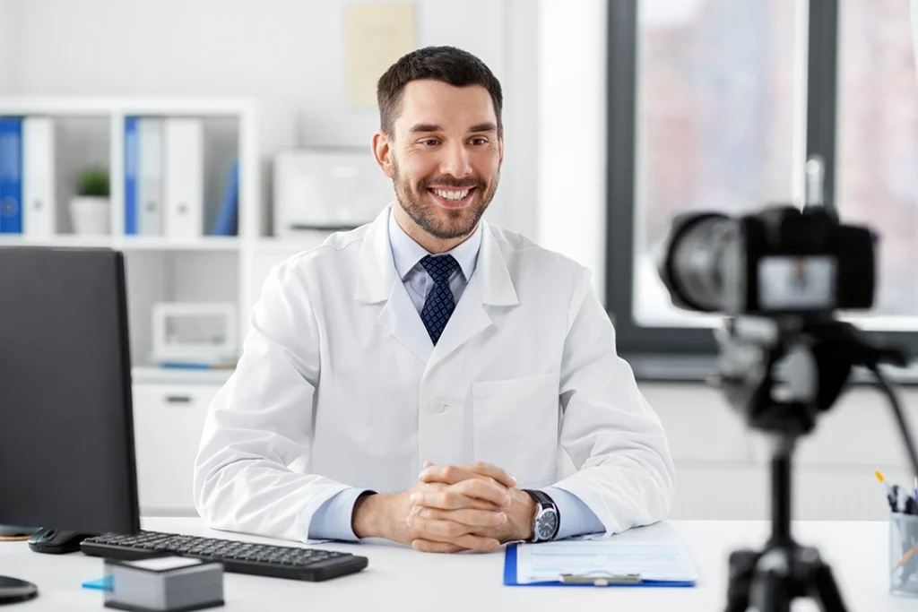 Dental videos company Sugar Land: Confident doctor in lab coat with hands clasped speaking directly to camera in modern clinic, delivering informative dental video with computer nearby.