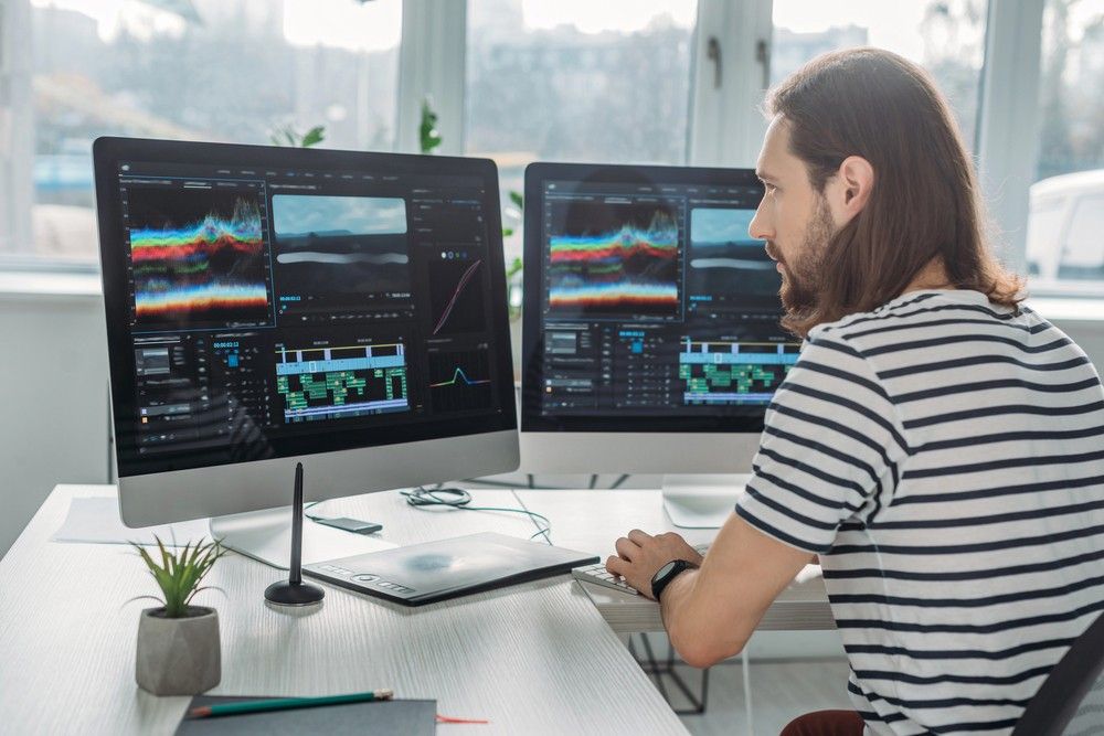 A man with long hair edits video footage on two large monitors for the corporate video production sugar land process.