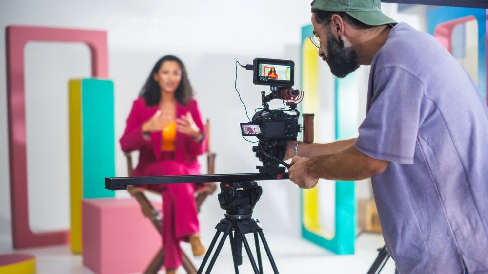Videographer from videography company Sugar Land captures an interview with a woman in a pink suit on a colorful set.